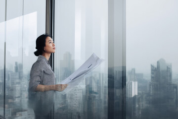 A middle-aged Asian woman reviews printed analytics in a sleek glass office, framed by a city skyline that hints at growth, ambition, and her leadership vision.