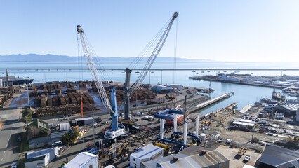 Nelson, New Zealand - 18 June 2025: Aerial view of the port, where towering cranes dominate the landscape, casting long shadows over the bustling docks filled with timber and vessels.