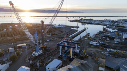 Nelson, New Zealand - 18 June 2025: Aerial view of the port bathed in the soft glow of sunrise, cranes towering over structures, silhouetted against the water and distant hills.