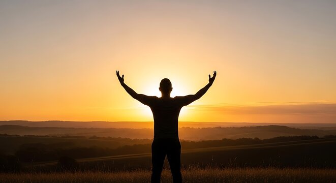Silhouette of a man with arms raised towards the sun during sunset, overlooking a landscape with rolling hills.