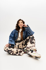 Stylish young woman lounging in cow print pants and denim jacket against a grey backdrop