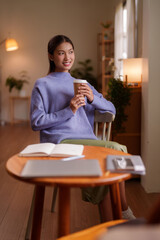 Young Asian woman enjoys morning coffee at home relaxing in cozy living room planning future tasks