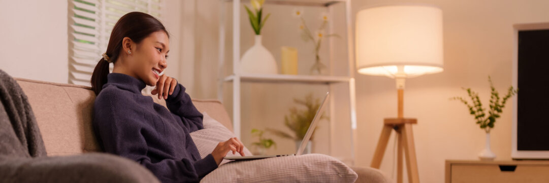 Asian adult woman smiling remote working online business productive tasks using laptop computer cozy home living room environment modern lifestyle digital connectivity comfort at home