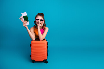 Smiling young woman posing with orange suitcase and travel essentials against a blue background,...