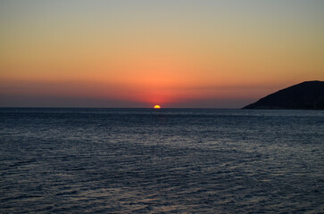 The ocean is calm and the sky is a beautiful orange color. The sun is setting, creating a warm and...