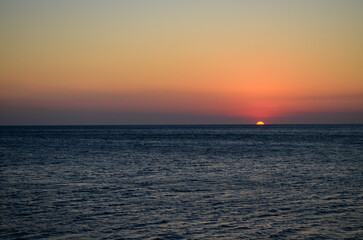 The ocean is calm and the sky is a beautiful orange color. The sun is setting, creating a warm and...