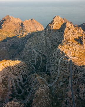 Aerial view of a winding road snaking through the rugged, sun-kissed mountains of Nus de sa Corbata, where shadows dance with the golden peaks, Escorca, Mallorca, Spain.
