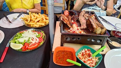 Traditional Argentinian barbecue, a parrillada with grilled meats like ribs and sausage, served on a mini charcoal grill. Accompanied by french fries, fresh salad, and chimichurri sauce.