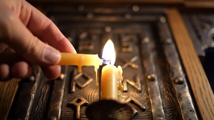 A hand lights a beeswax candle with another, over a metal plate adorned with runes