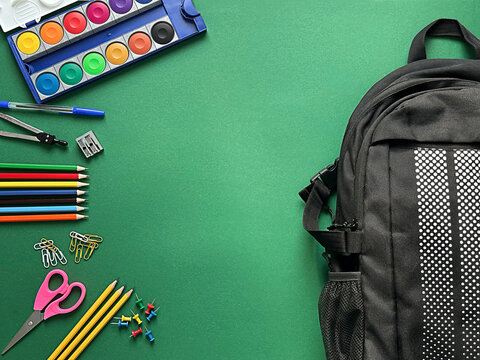 A textured green background with a black backpack and various school supplies including colored pencils, scissors,