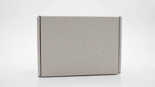 A plain, light gray cardboard box stands on a white surface, angled slightly towards the viewer. The box has visible seams and texture