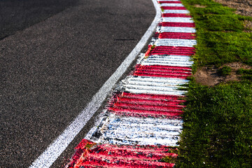 Detail view of a red and white painted curb on a racing track, with adjacent asphalt and green grass. Motorsport infrastructure and track environment