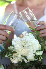 The newlyweds raise a toast with glasses of white sparkling champagne against the background of wedding rings and a wedding bouquet.