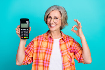 Smiling senior woman holding card terminal and making OK hand gesture against teal background