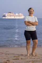 Tourist smiling with arms crossed on beach at dusk with cruise ship in background