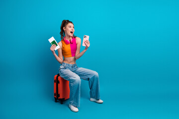 Happy young woman sitting on a suitcase with a passport and phone, ready for travel against a...