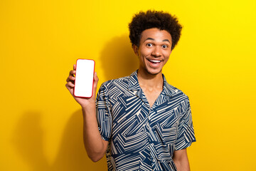 Happy young man in fashionable shirt showing smartphone against vibrant yellow background,...