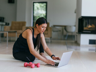 A young woman in sportswear is sitting in the living room and preparing for online training while using a laptop.