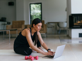 A young woman in sportswear is sitting in the living room and preparing for online training while using a laptop.