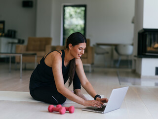 A young woman in sportswear is sitting in the living room and preparing for online training while using a laptop.
