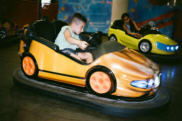 A young boy drives an orange bumper car with focus and excitement, while a girl follows in a yellow car, captured in a candid 35mm film photo at an indoor amusement park.