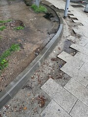 Broken pavement and eroded sidewalk in urban area after rainfall. Poor drainage and street infrastructure problems visible