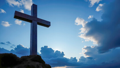 A large wooden cross sits on rocky ground at sunset against a dramatic backdrop of clouds. Silhouetted in soft twilight light