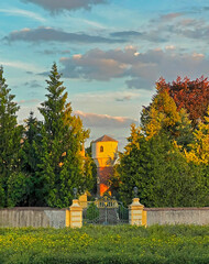  Park still life with a historic brick water tower at sunset. Behind the flowerbed, a metal gate and walls with summer July trees in autumn colors. Europe.