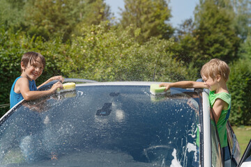Two boys are washing the windshield of a car. They are rubbing the glass with sponges.