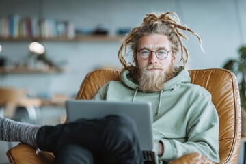 Man with dreadlocks sits in chair at home and works on a laptop, focusing with a serious look, wearing a hoodie and round spectacles, looking at the camera.