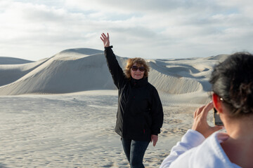Cheerful senior woman greeting, standing amid pristine sand dunes beneath radiant blue sky during outdoor photography session