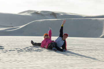 Tourists sliding down white sand dune, arms waving with joy in expansive desert environment