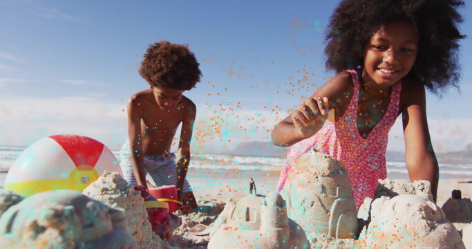 Siblings in pink dress and trunks building sandcastles on beach with red bucket, plastic shovel