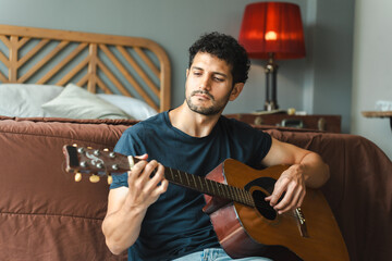 Young man sitting on bedroom floor playing acoustic guitar, gazing thoughtfully into distance,...