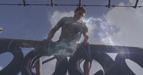 Climbing man wearing grey t-shirt black shorts gripping beam over strapped tires on obstacle course