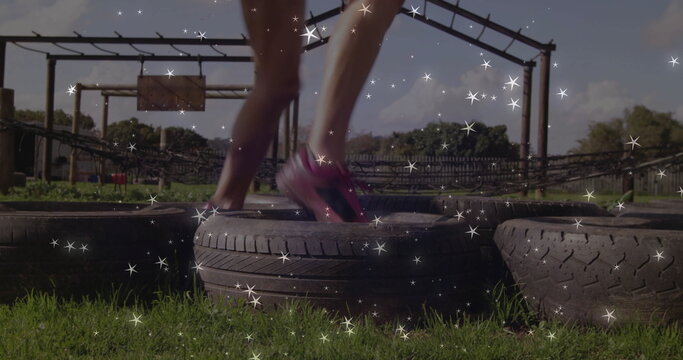 Hopping woman in pink running shoes moving through tires on grass obstacle course, with rope nets