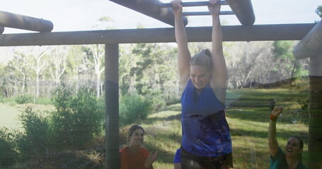 Hanging woman in blue tank top gripping wooden monkey bars in park, with teammates cheering