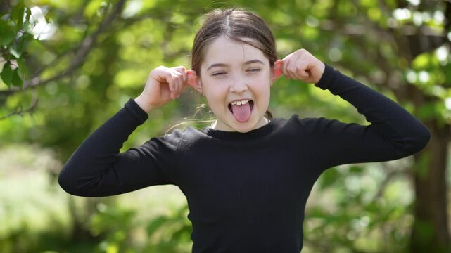 Teenage girl goofing around, pulling her ears to sides and pretending to be monkey, having fun in park. Young adolescent playing playfully, stretching her ears and mimicking monkey while enjoying