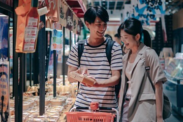 Young couple browses pre‑packed fresh foods in a bright supermarket, examining items together and...