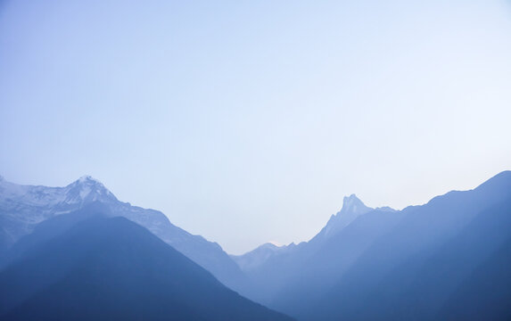 Snow-capped Mount Annapurna peaks visible in the morning with ray of light and mist at Annapurna Base Camp, Nepal with blue sky and white clouds background.