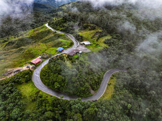 A breathtaking aerial view of a winding mountain road cutting through lush green forest, partially veiled by patches of low-hanging clouds. 