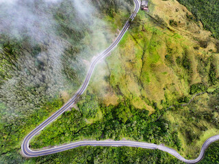 A breathtaking aerial view of a winding mountain road cutting through lush green forest, partially veiled by patches of low-hanging clouds. 