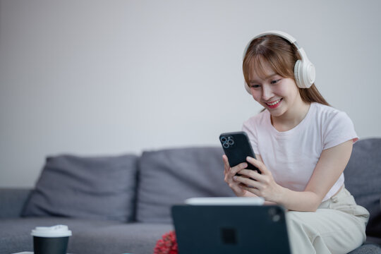Young Asian woman using smartphone and listening music with headphones while sitting on comfortable sofa in living room at home, she is relaxing on weekend