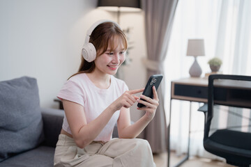 Happy asian woman wearing wireless headphones sitting on comfortable sofa and choosing playlist...
