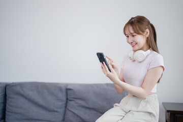 Happy young woman sitting on the edge of a grey sofa using her smartphone with headphones around...