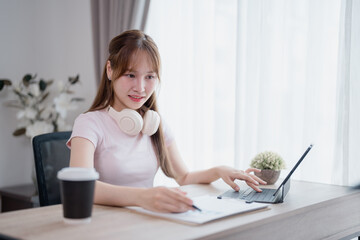 Young asian woman with headphones around her neck is working from home, using a tablet and taking...