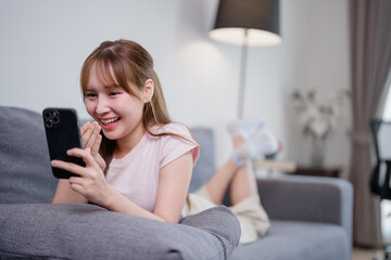 Happy Asian woman laughing and using her smartphone while lying on the sofa in a cozy living room,...