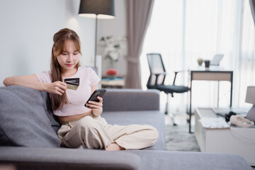 Asian woman sitting on sofa using smartphone and credit card for online shopping at home, she is...