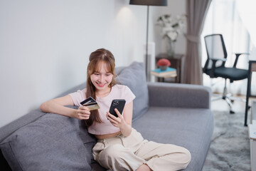 Obraz premium Young Asian woman relaxing on a comfortable sofa, holding a credit card while using a smartphone for online shopping in her cozy living room, enjoying the convenience of e-commerce