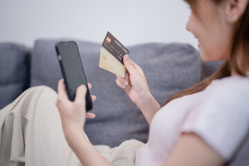 Woman sitting comfortably on a sofa at home, holding a credit card while using a smartphone for...